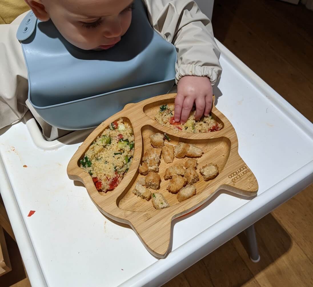 Brandon eating couscous with veg and not-chicken nuggets from his bamboo plate, sitting in his high chair